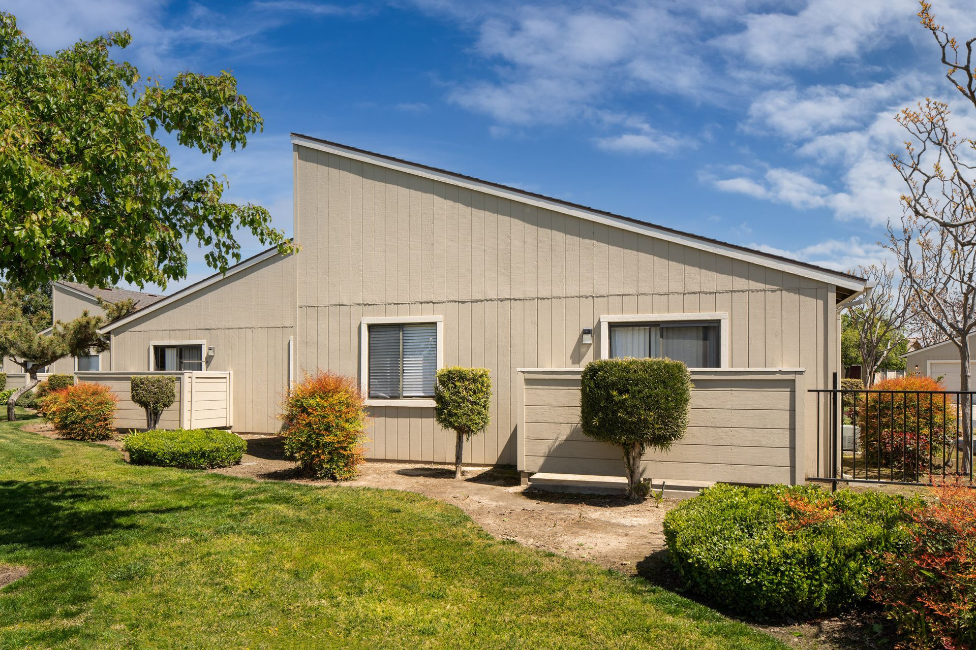 Tan, single-story house with angled roof, windows, and manicured landscaping under a blue sky.
