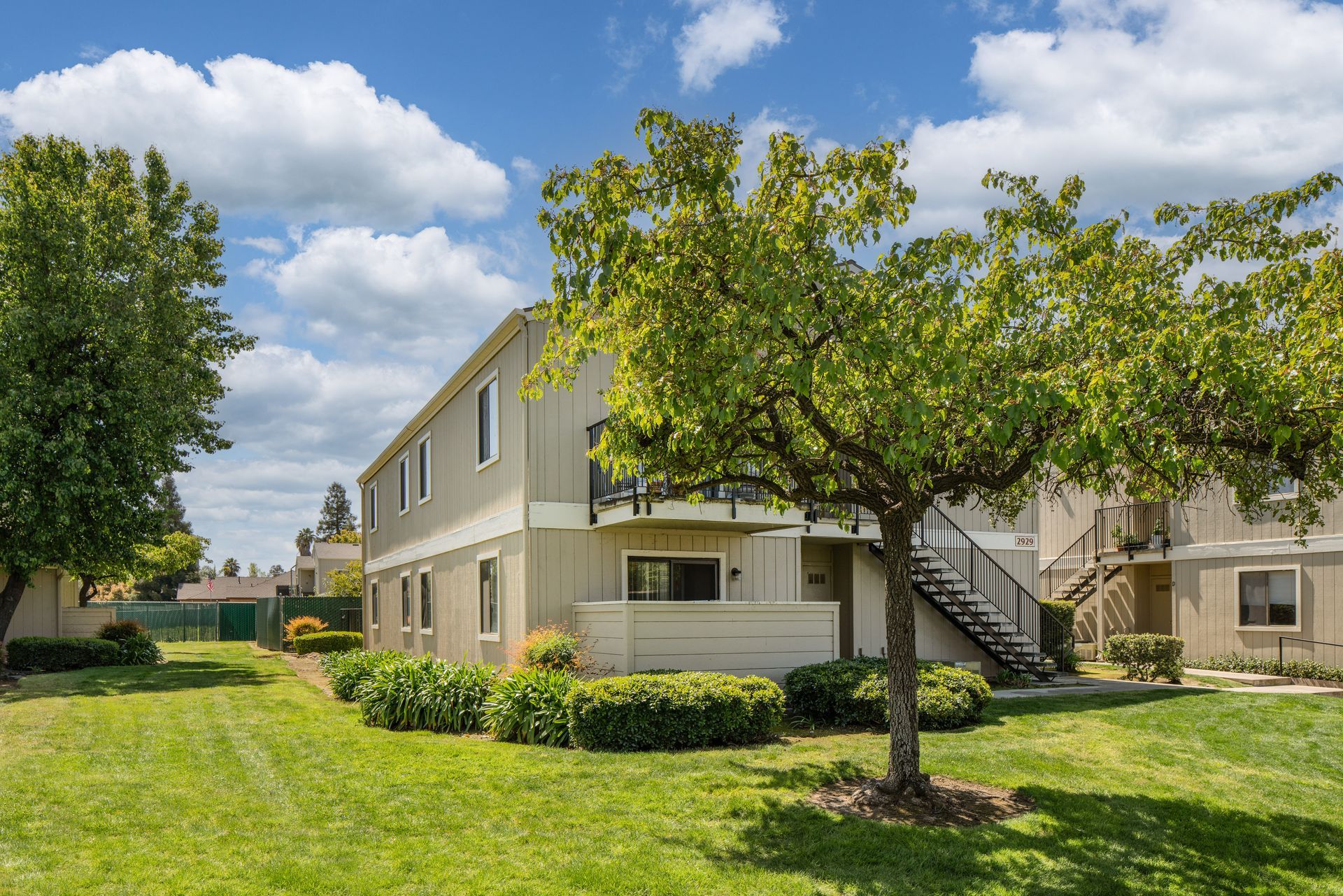 Two-story apartment building with tan siding, surrounded by green grass and trees, under a cloudy blue sky.