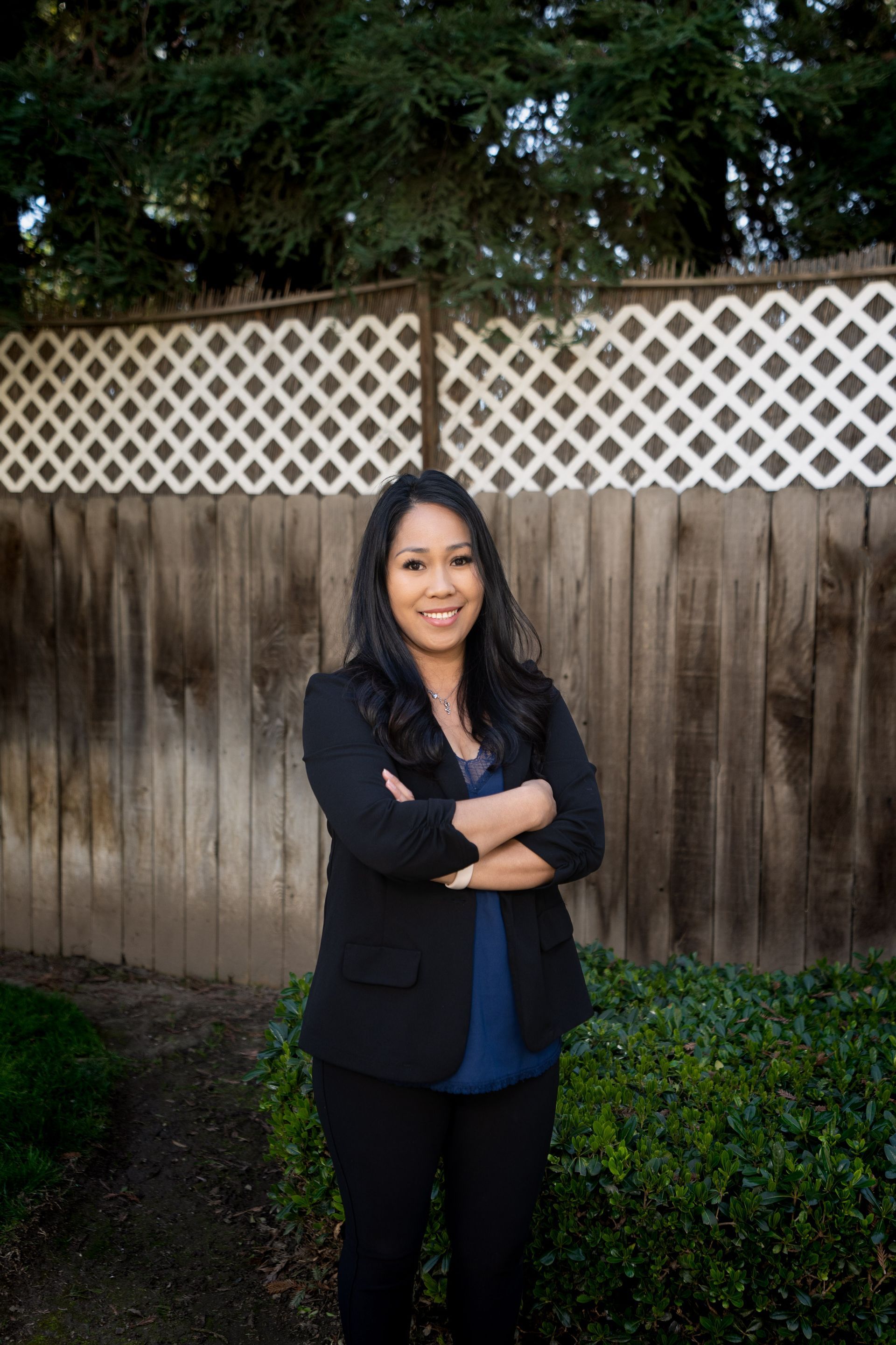 Woman with crossed arms, smiling in front of a fence and foliage.
