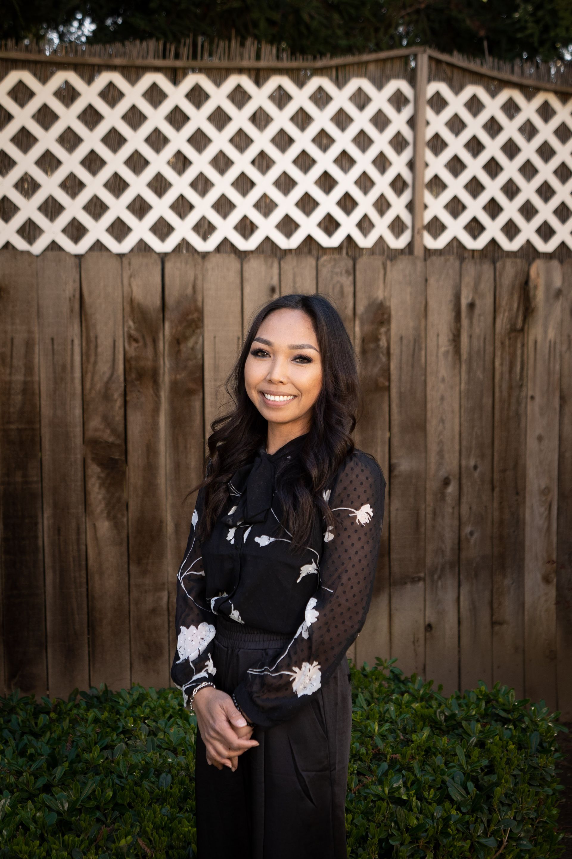 Woman with dark hair smiles, wearing black dress with floral pattern, standing in front of wooden fence with white trellis.