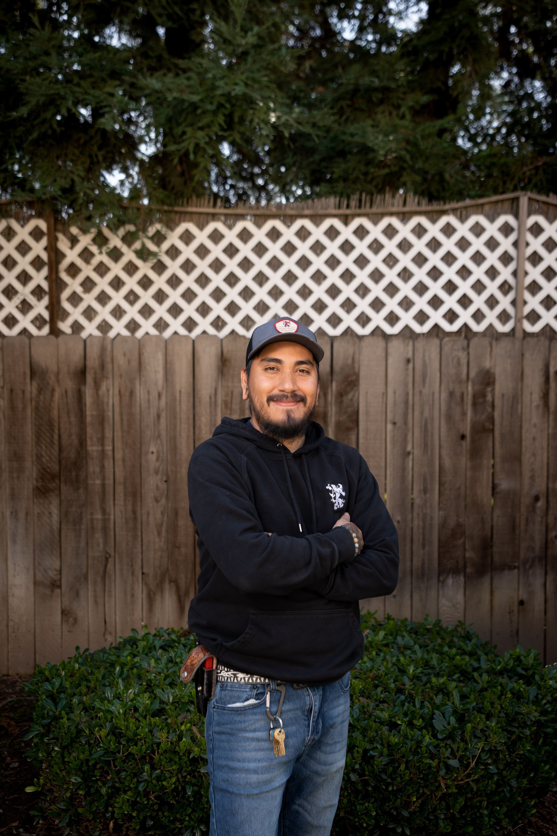 Man with crossed arms standing in front of a fence, wearing a hat, hoodie, and jeans.