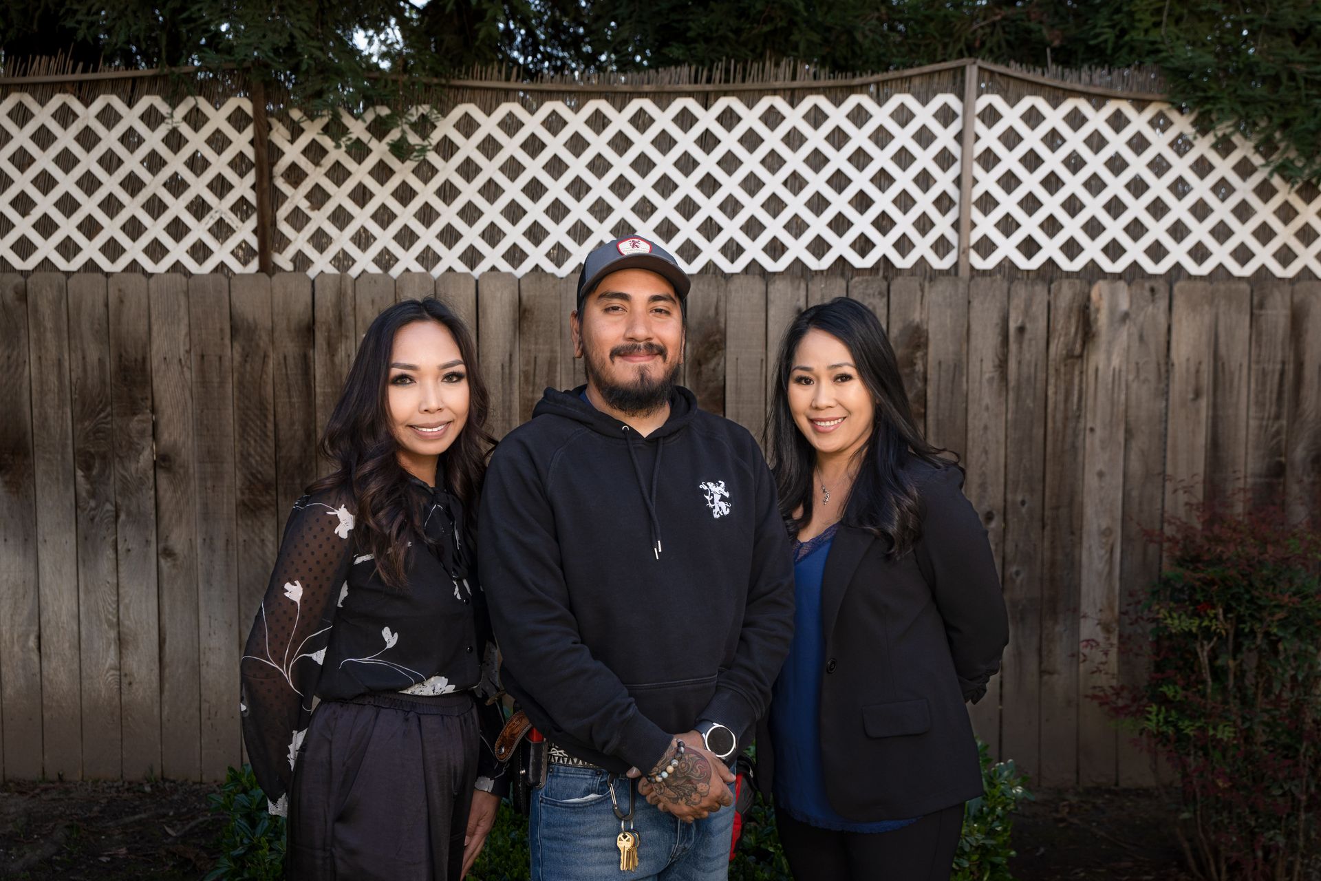 Three people pose in front of a fence. A man stands between two women, all smiling at the camera.