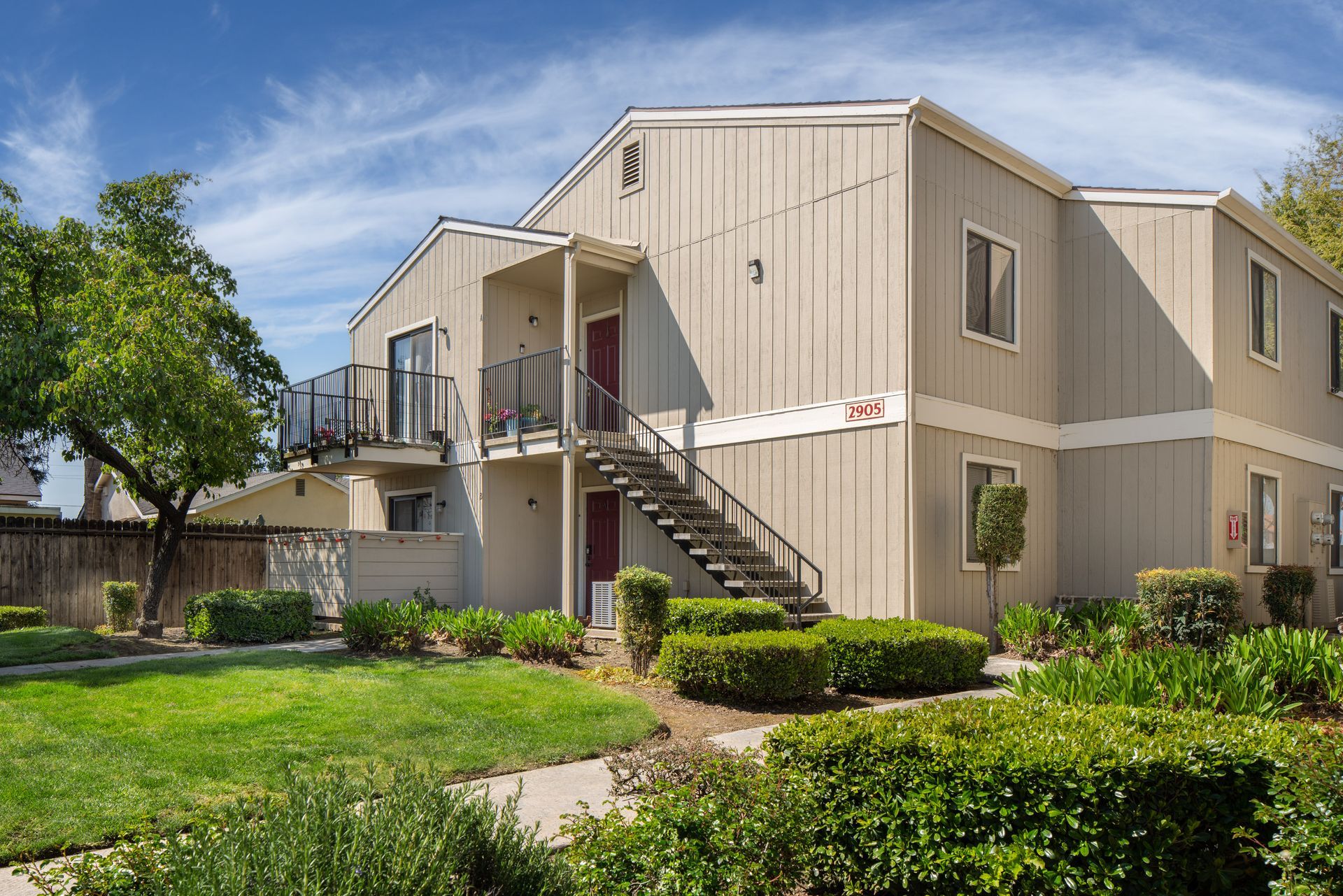 Two-story beige apartment building with exterior stairs, surrounded by green grass, bushes, and trees.