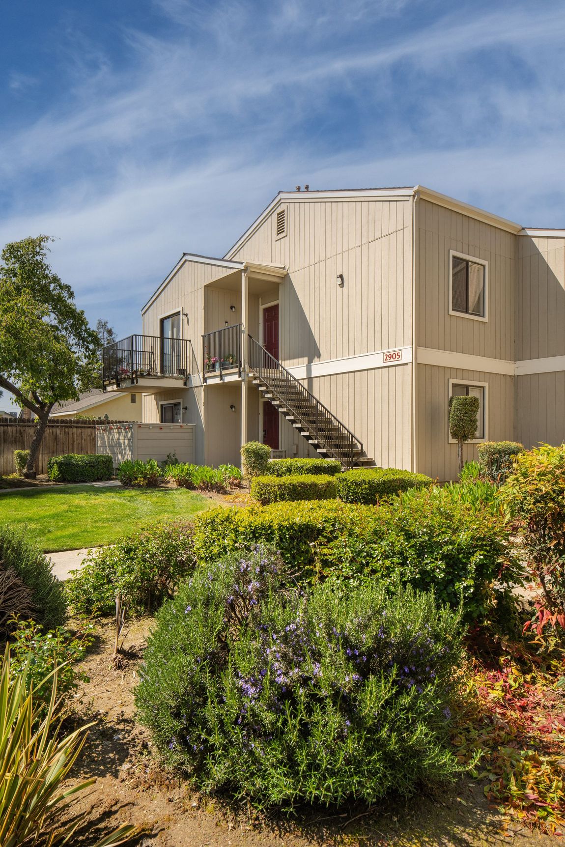 Two-story beige apartment building with outdoor stairs, surrounded by green shrubs and blue sky.