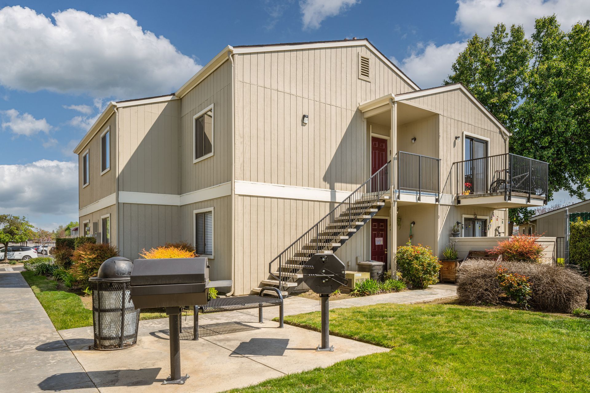 Two-story beige apartment building with stairs, grill, and landscaping. Sunny day.