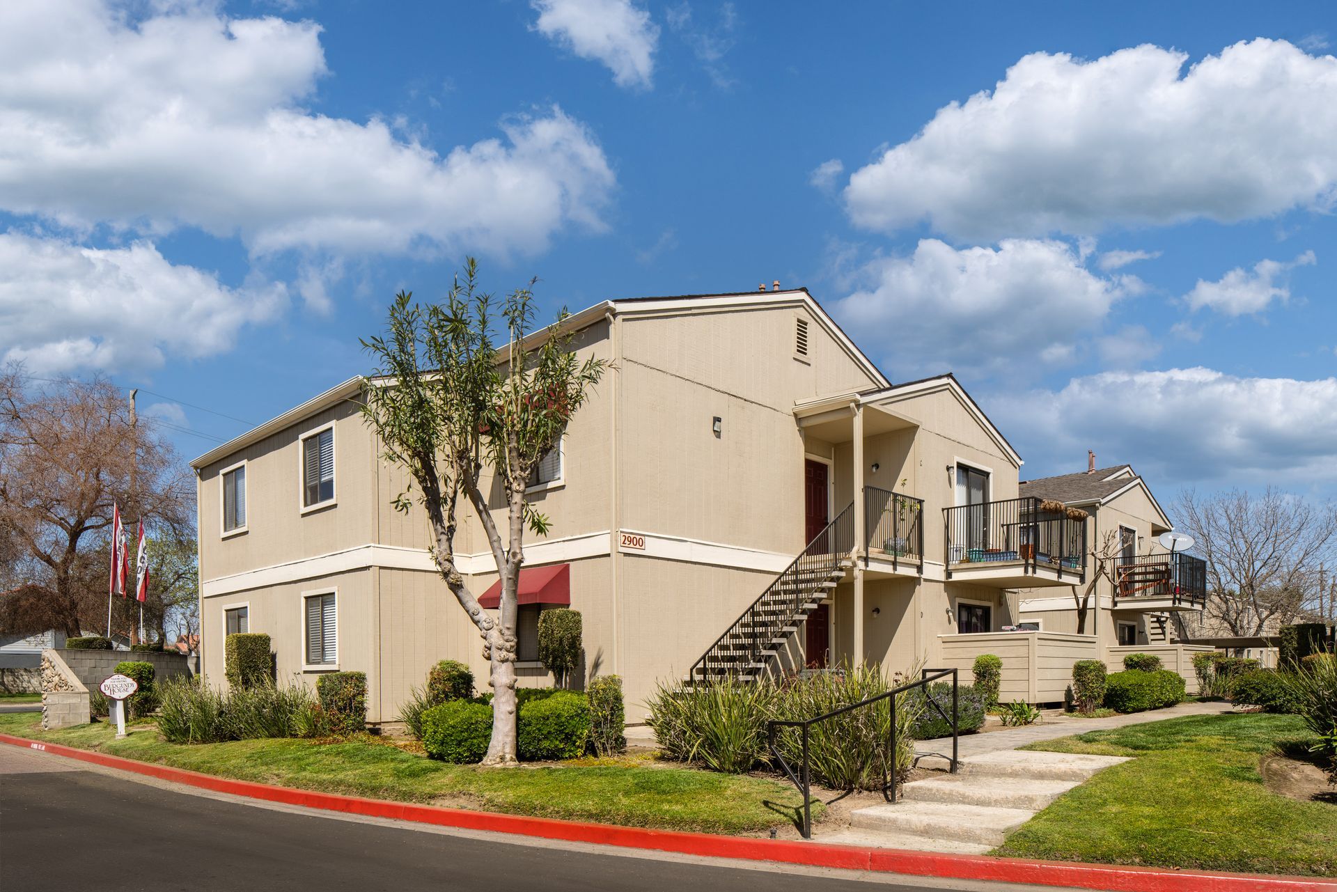 Two-story beige apartment building with stairs and small balconies; blue sky, green grass and bushes.
