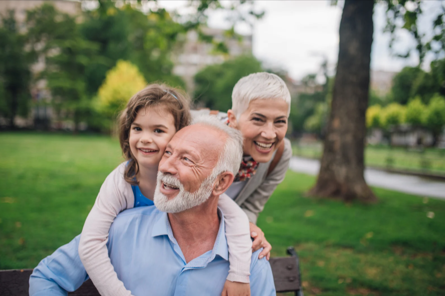 Two grandparents playing with their granddaughter in a park, smiling and enjoying the outdoors together.