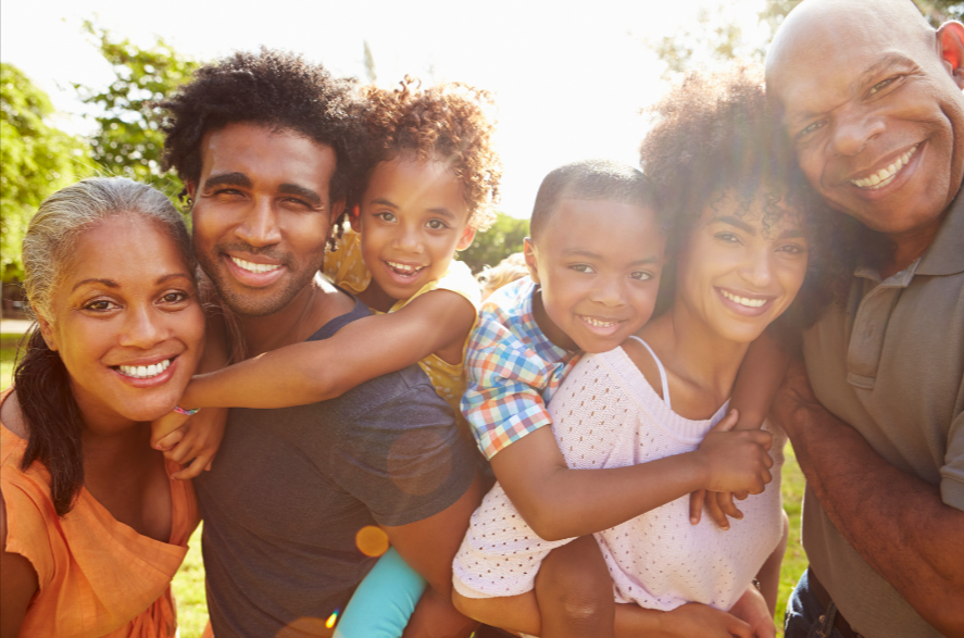 A multigenerational African American family posing for a picture in a park. The two grandchildren, a boy and a girl, are playfully hanging on the backs of their parents.