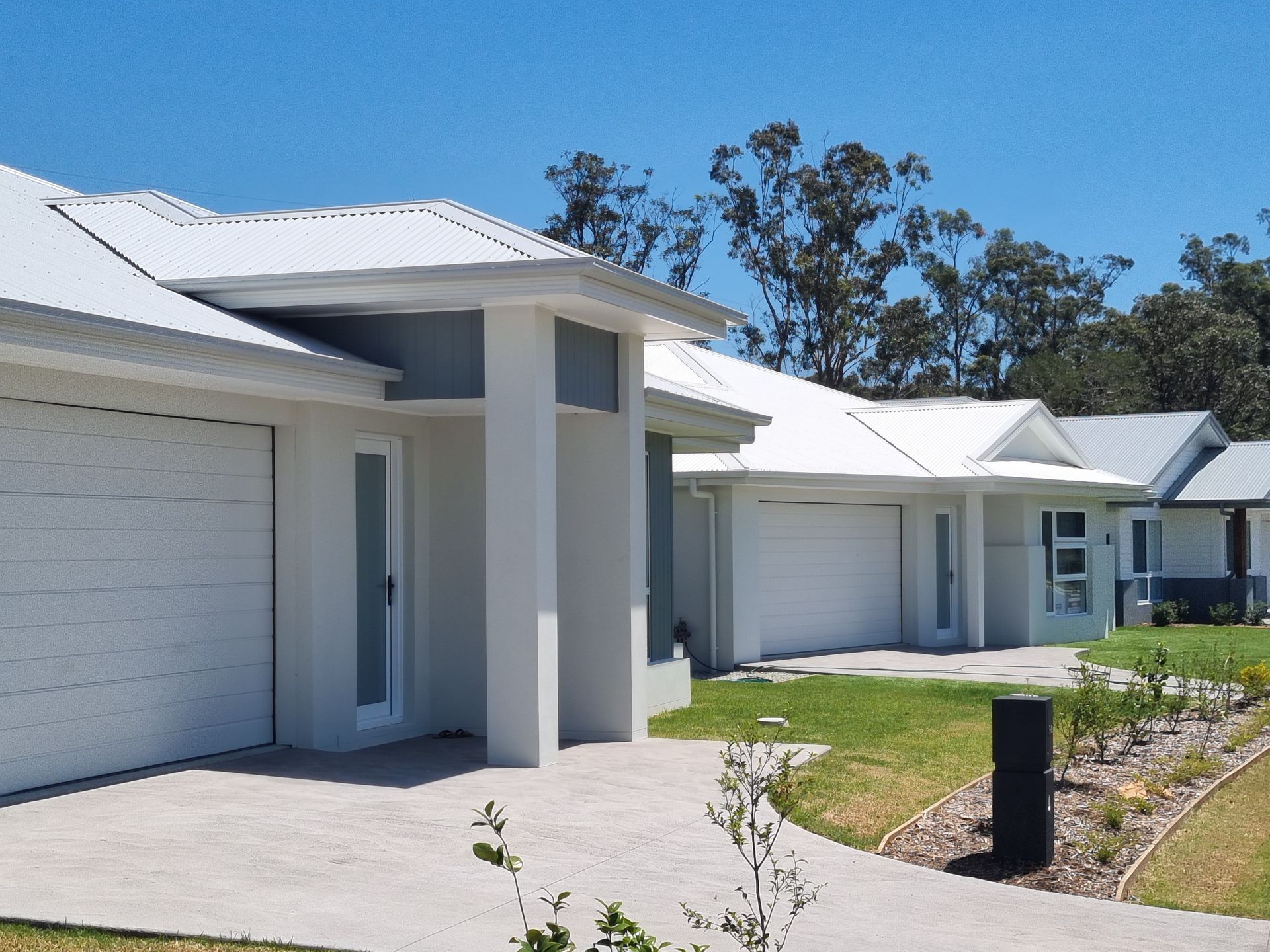 Row of modern, single-story homes with white roofs, gray walls, and attached garages, set on green lawns under a blue sky.