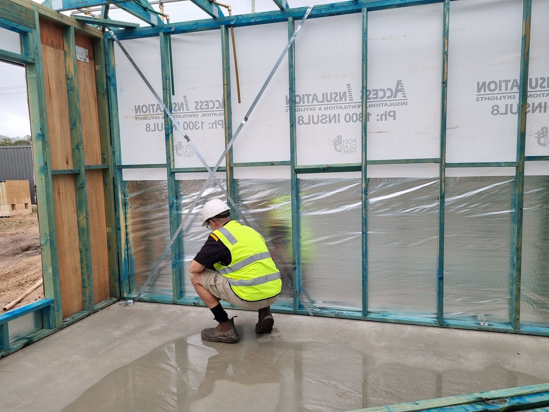 Construction worker in a reflective vest squats in a room with wet concrete floor and wood framing.