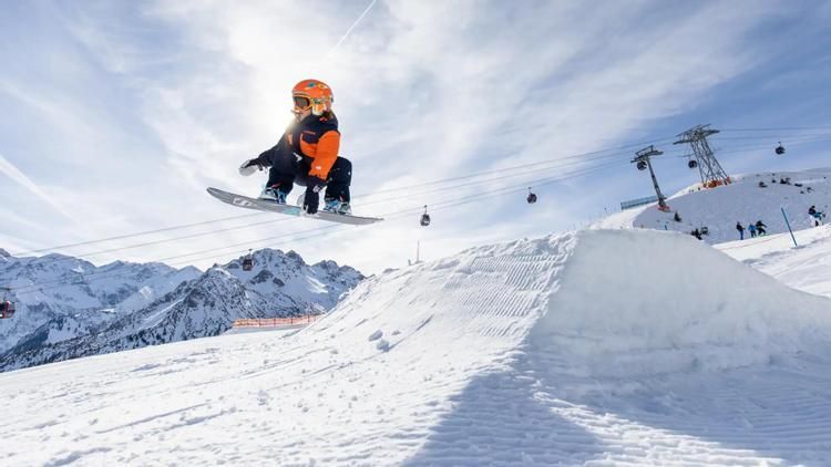 Snowboarder springt von einer Schneerampe in einem Skigebiet mit Bergen und blauem Himmel.