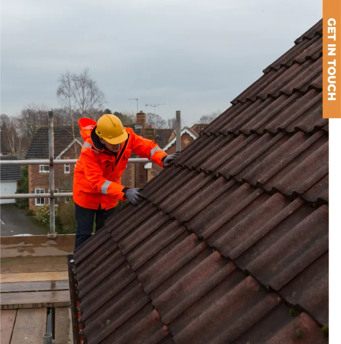 A man in an orange jacket and yellow hard hat is working on a roof