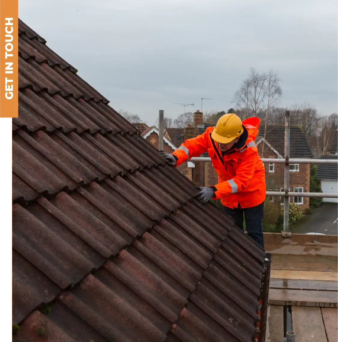 A man wearing an orange jacket and yellow hard hat is working on a roof