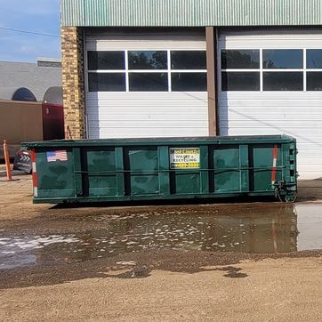Green dumpster in front of a building with garage doors. Puddle of water in the foreground.