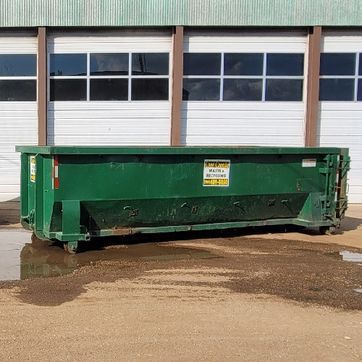 Green dumpster in front of a white building with windows and brown beams, sitting in a puddle.
