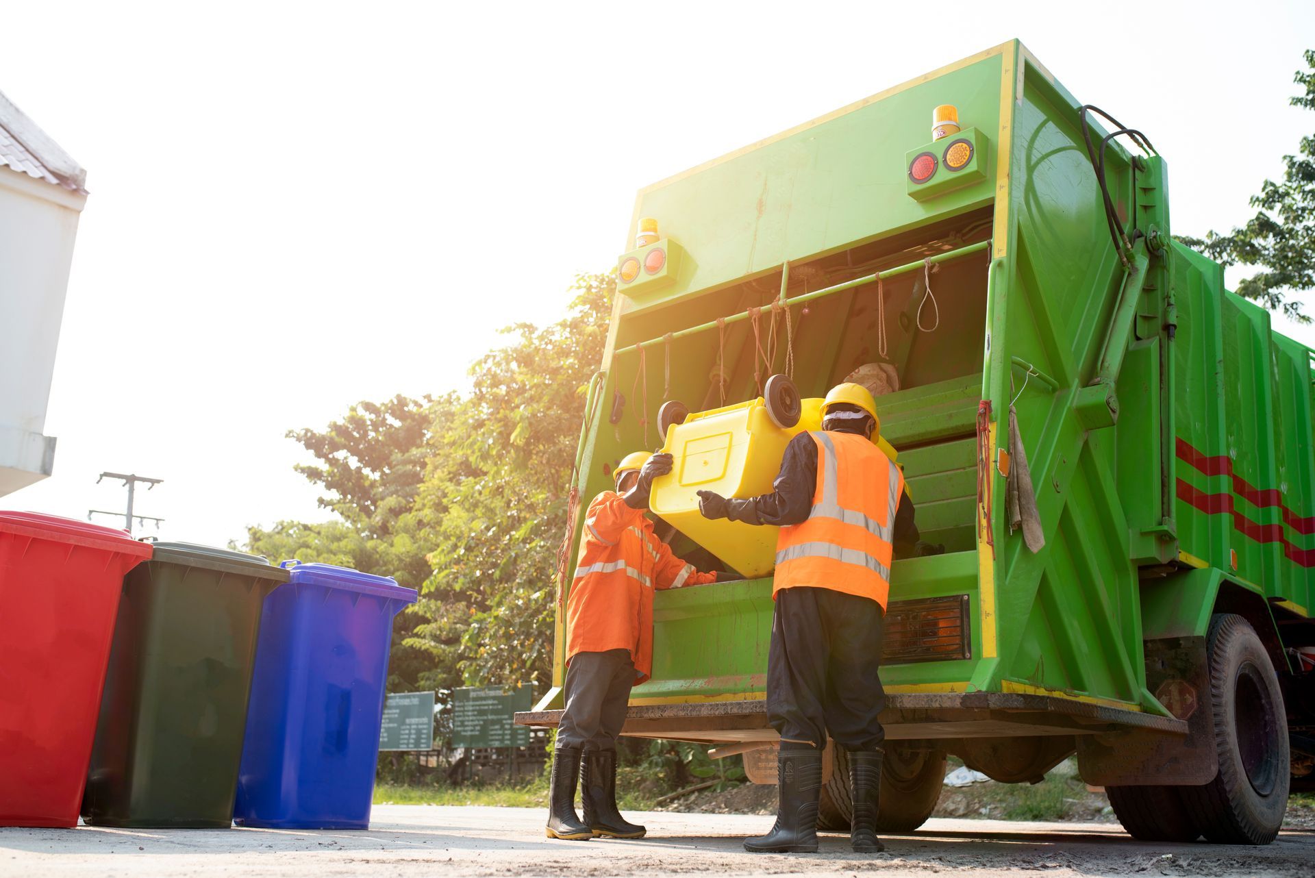 Waste collectors emptying a yellow bin into a green garbage truck near red, blue, and green bins under a bright sun.