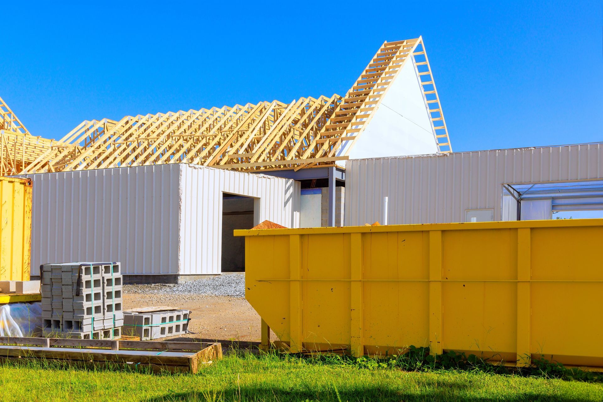 Construction site with wooden roof trusses, white siding, yellow dumpster, and blue sky.