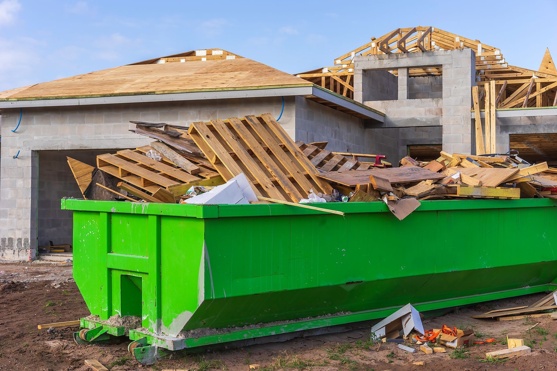 Green dumpster overflowing with construction debris in front of a house under construction.
