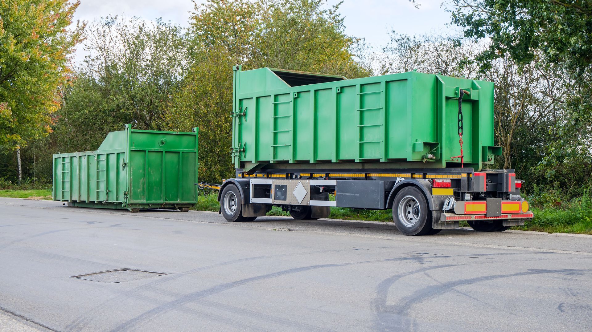 Green dumpster containers on a trailer parked on a road, trees in background.