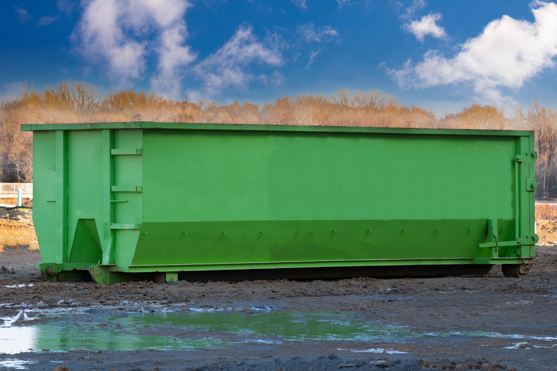 Green metal dumpster on dirt, trees and blue sky background.