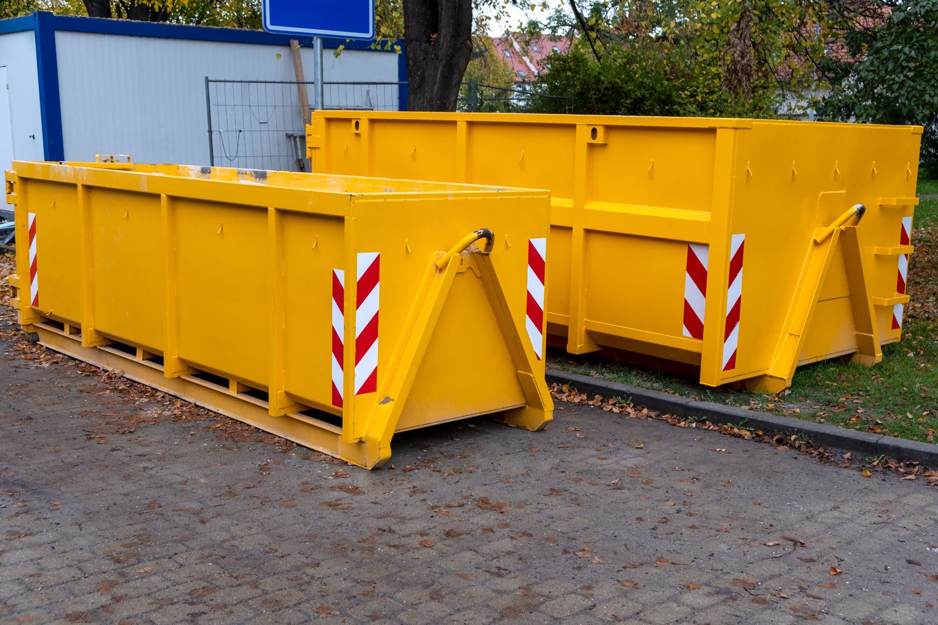 Two yellow dumpsters on a paved surface, with red and white diagonal stripes for visibility, outdoors.