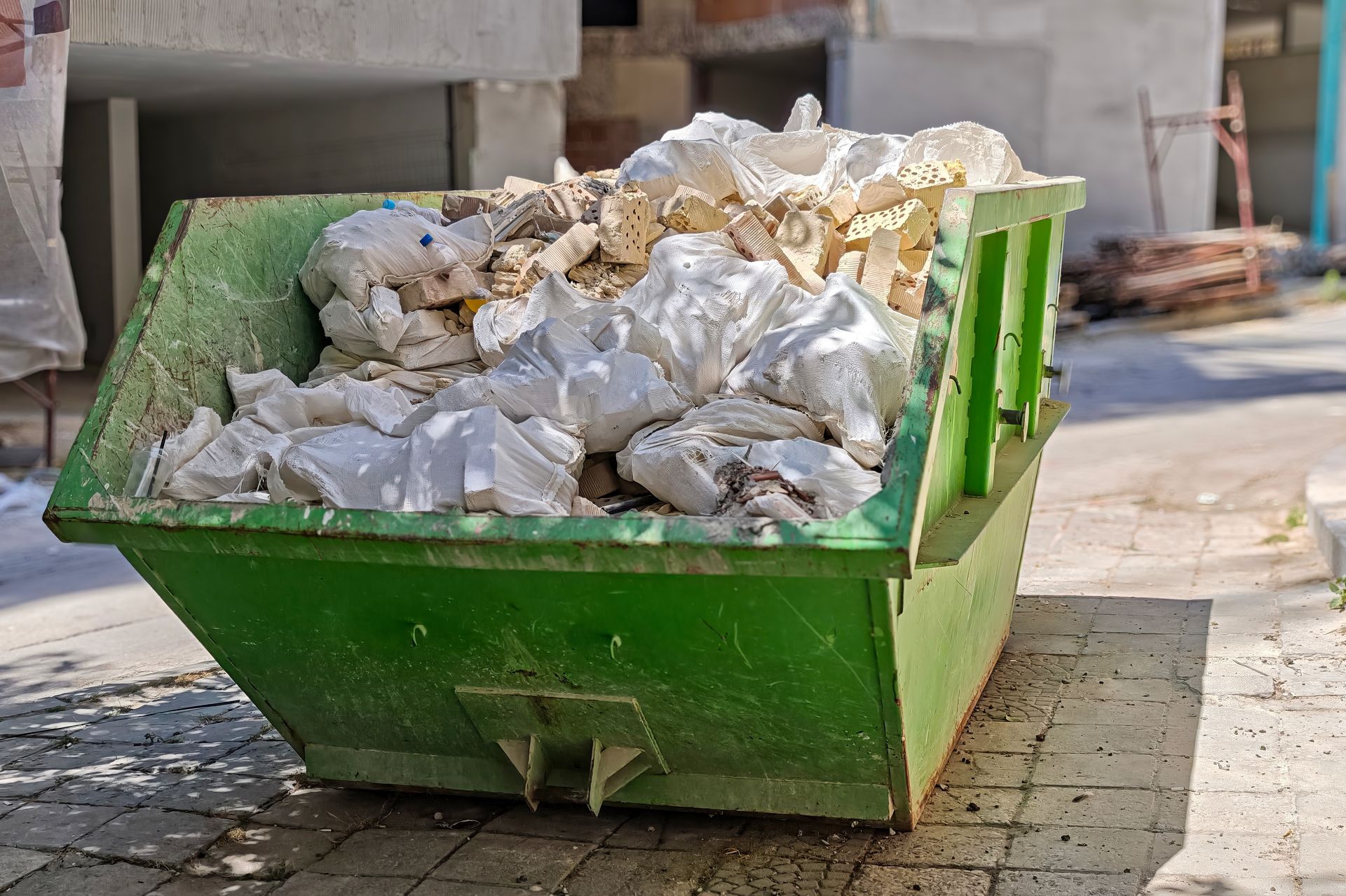 Green dumpster overflowing with construction debris on a sidewalk.
