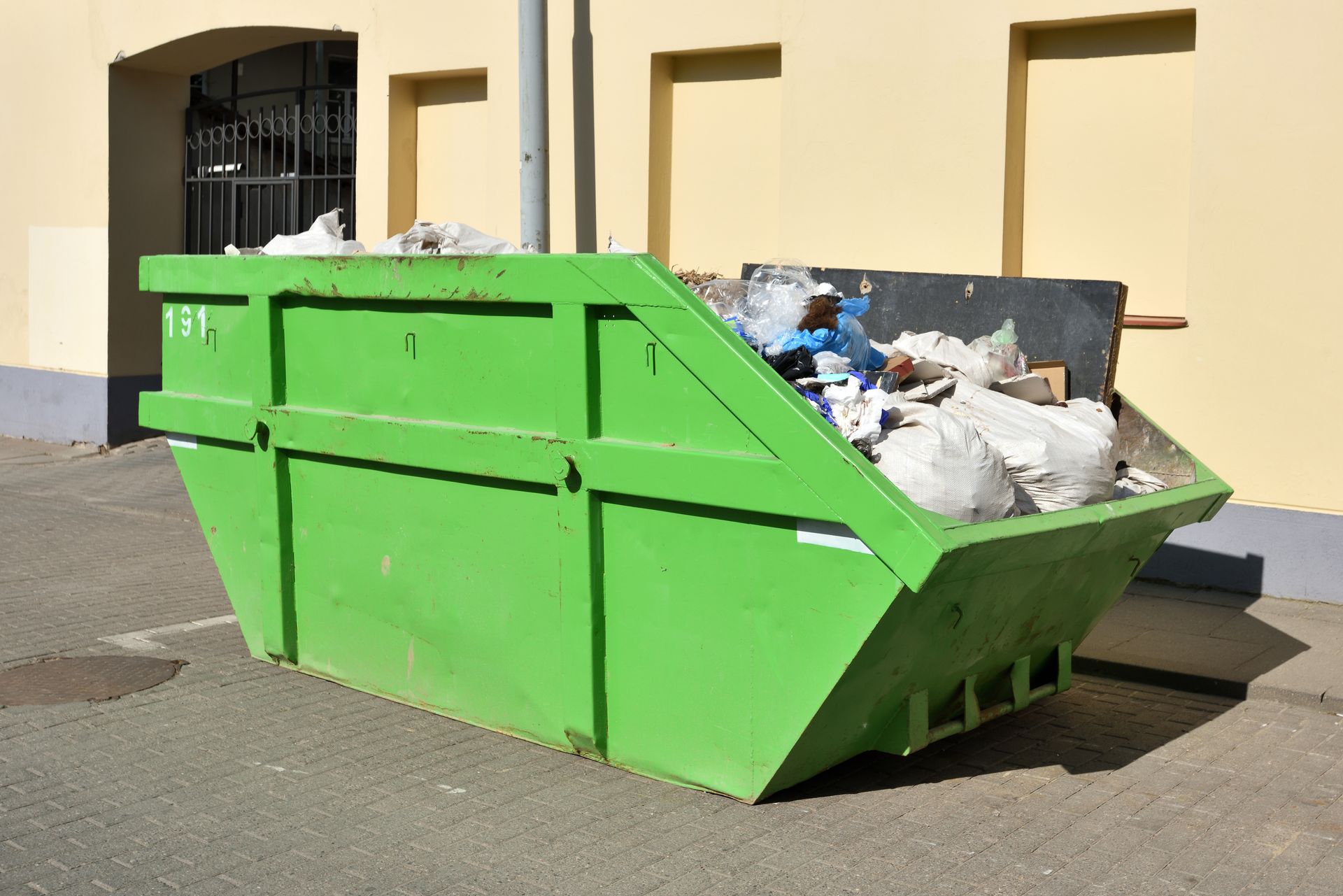 Green dumpster overflowing with trash, parked on concrete near a building.