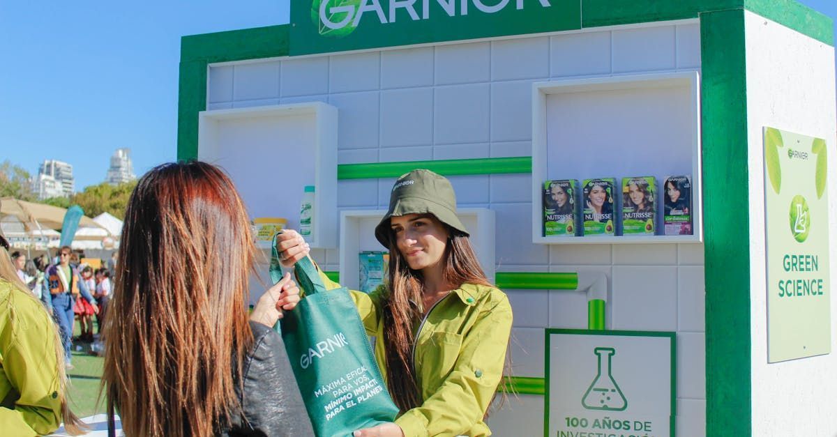 Woman hands a Garnier bag to another woman in a green setting, next to a branded display.