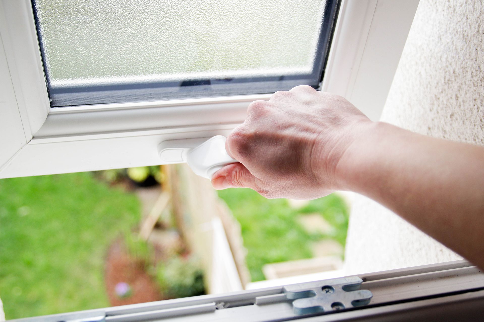 A hand gripping a white handle to open a double-glazed window with frosted glass and a garden view A hand gripping a white handle to open a double-glazed window with frosted glass and a garden view