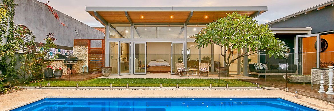 A house with a pool in its front yard. The house has glass walls. There is a tree in front of the house.