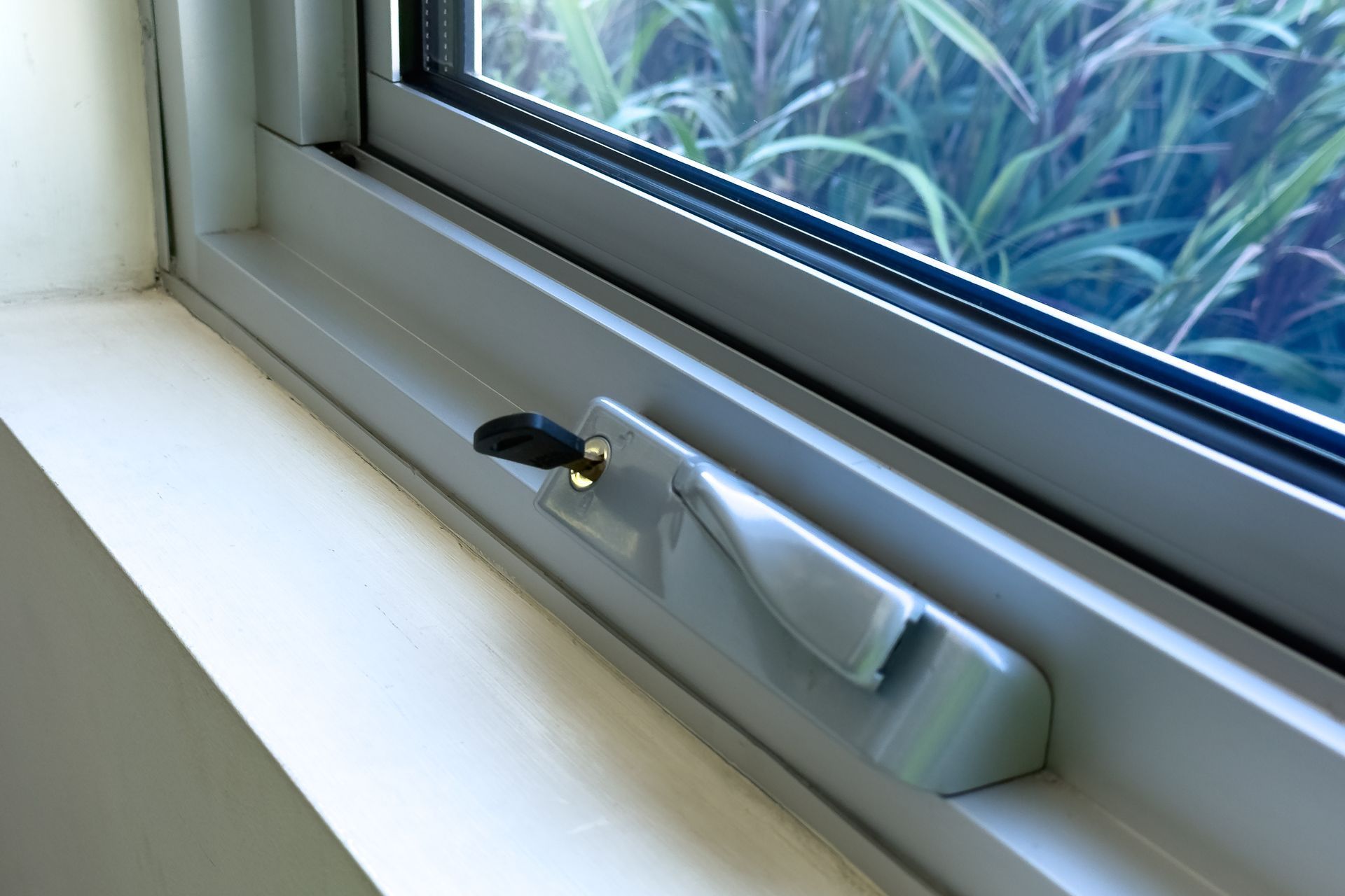 Close-up of a white window with a lever-style lock; window looks out to green foliage.