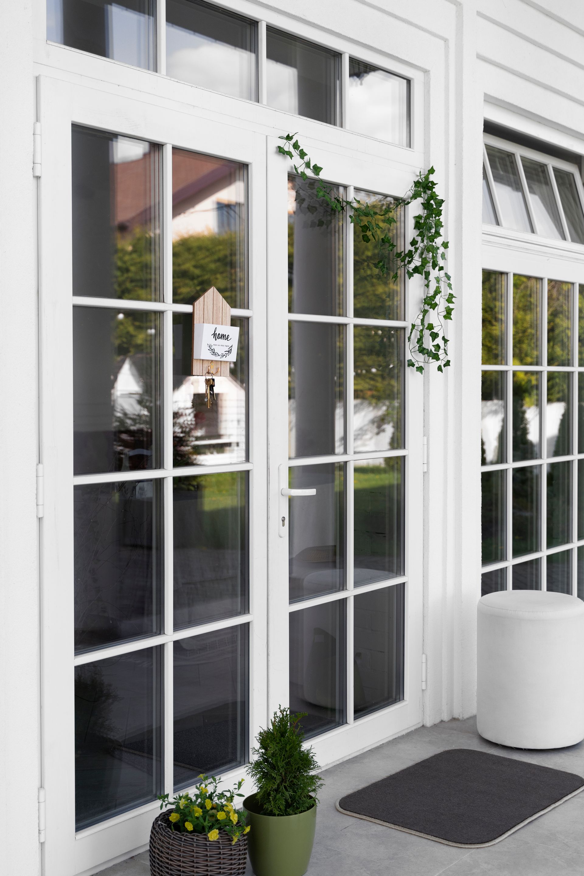 White French doors with glass panes, adorned with greenery and a small sign, are on a white porch.