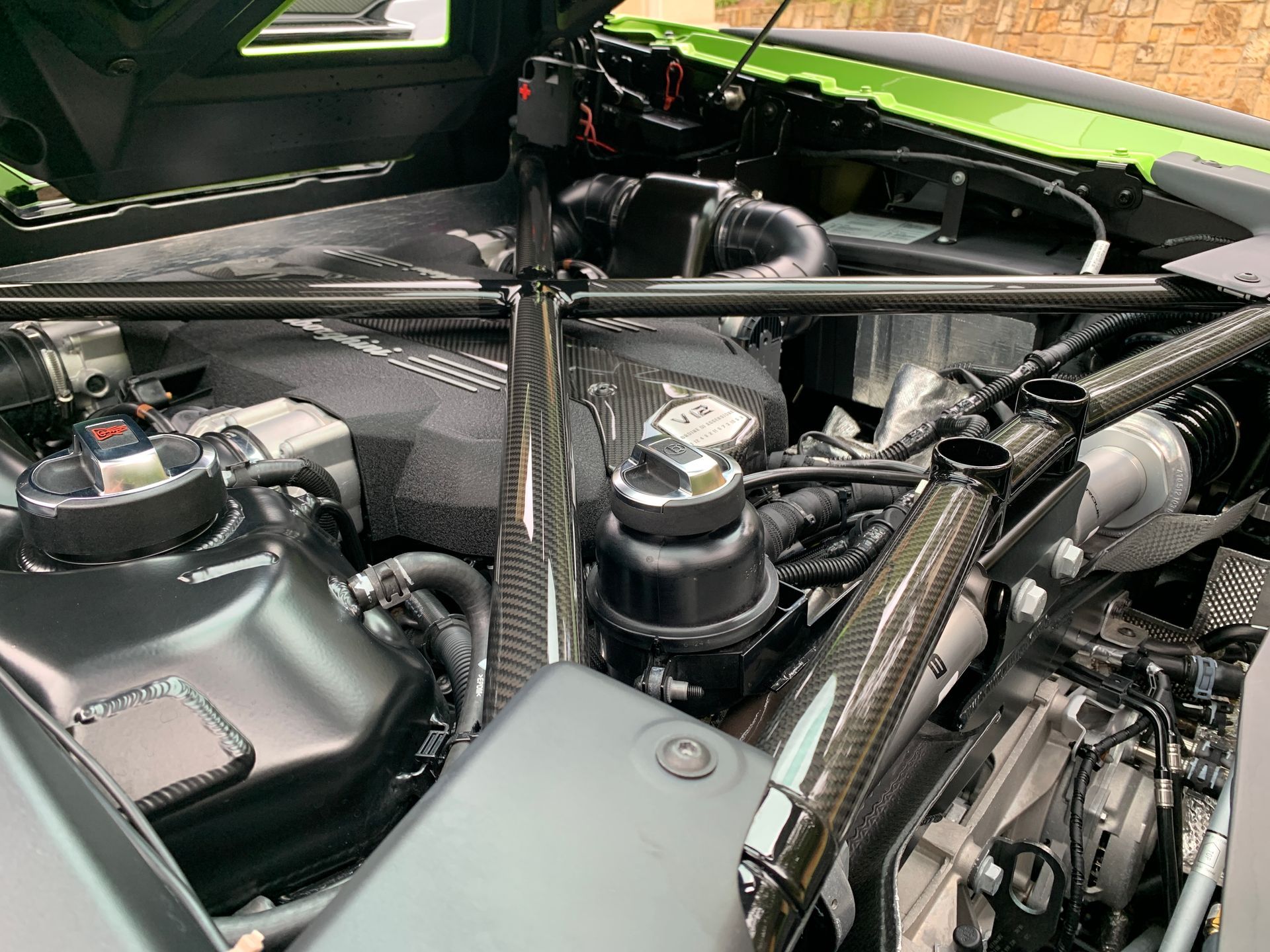 Lamborghini engine bay with carbon fiber accents. Green car exterior visible, various black components, and silver tubing.