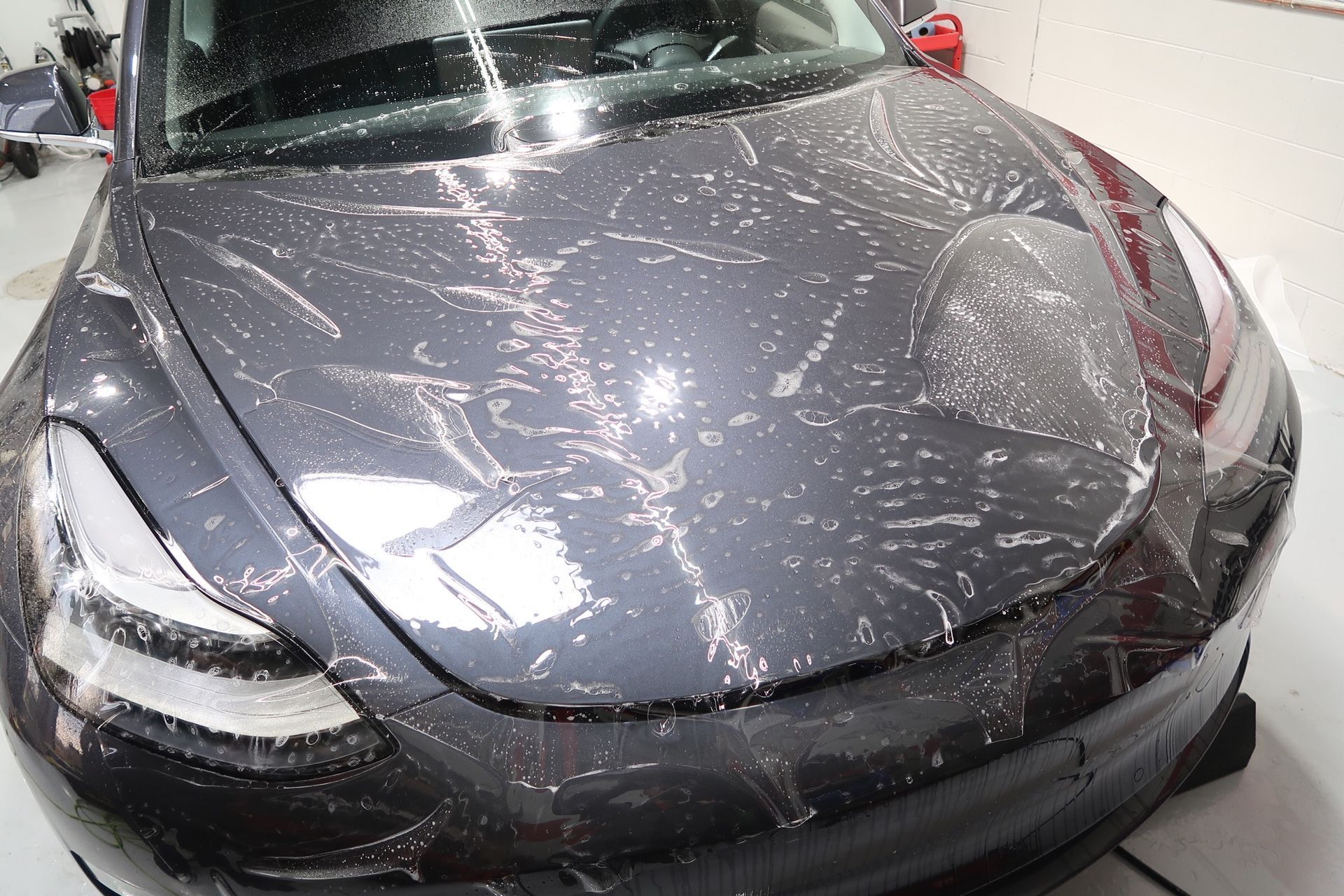 A gray Tesla car hood covered in protective film with water droplets.