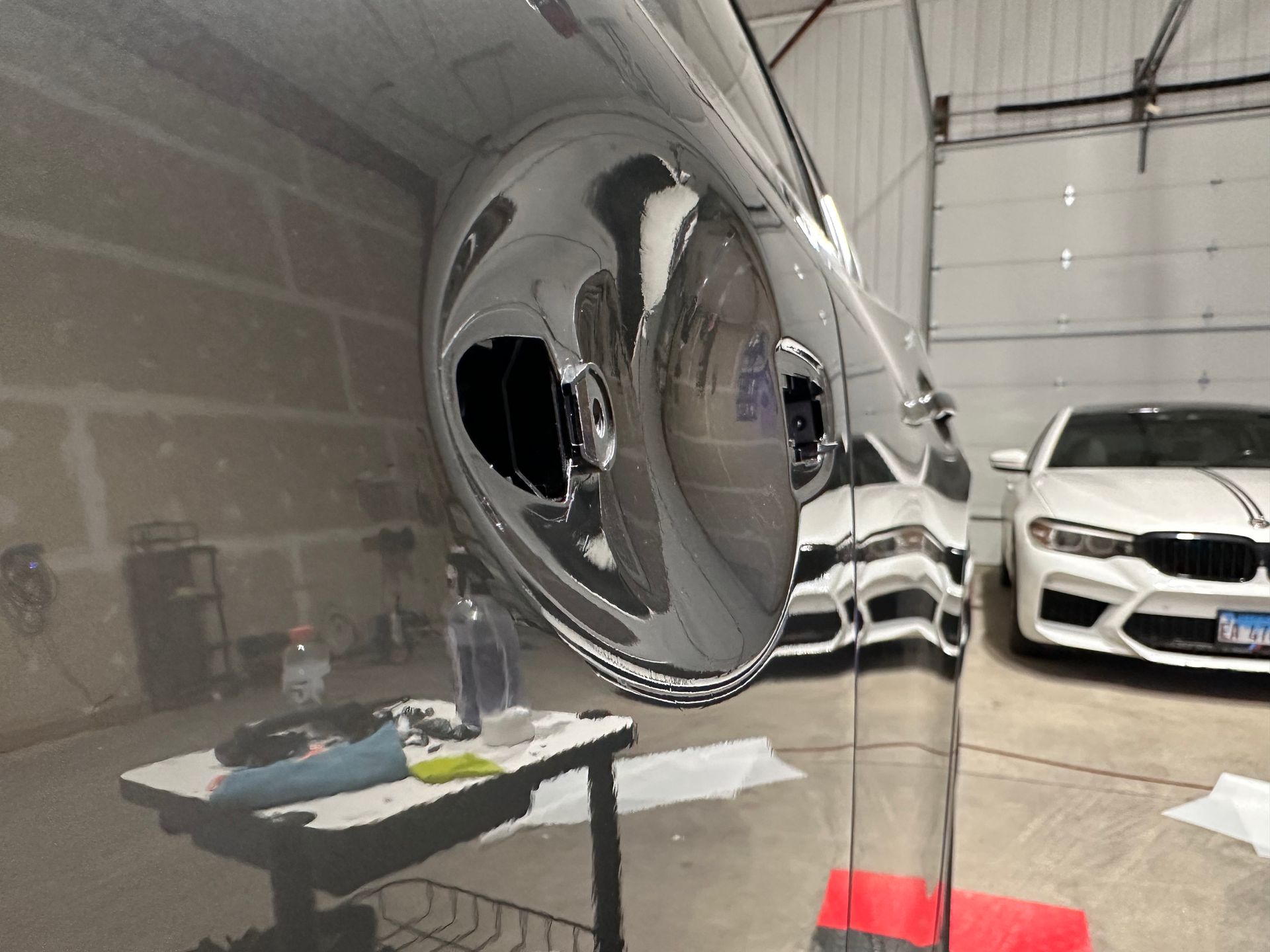Chrome car side mirror, garage setting; reflection of a white car, detailing supplies.