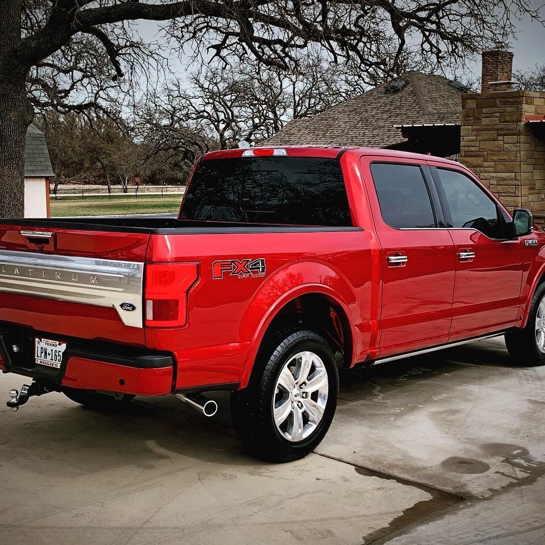 Red Ford F-150 pickup truck parked on a driveway in front of a stone building with a tree in the background.