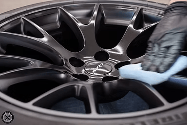 Black-gloved hand wipes down a shiny, dark gray car wheel with a light blue cloth.