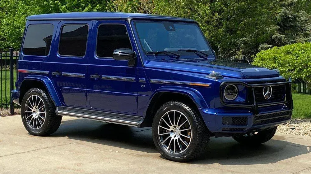 Blue Mercedes-Benz G-Wagon SUV parked on a paved driveway with green foliage in the background.