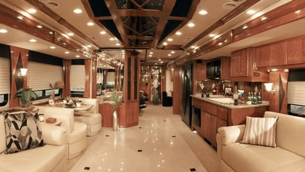 Interior of a luxury RV with wood paneling, cream sofas, and a kitchen, showcasing a comfortable living space.