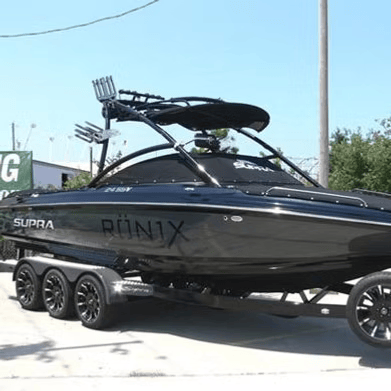 Black Supra Ronix wake boat on trailer, parked outside, sunny day.