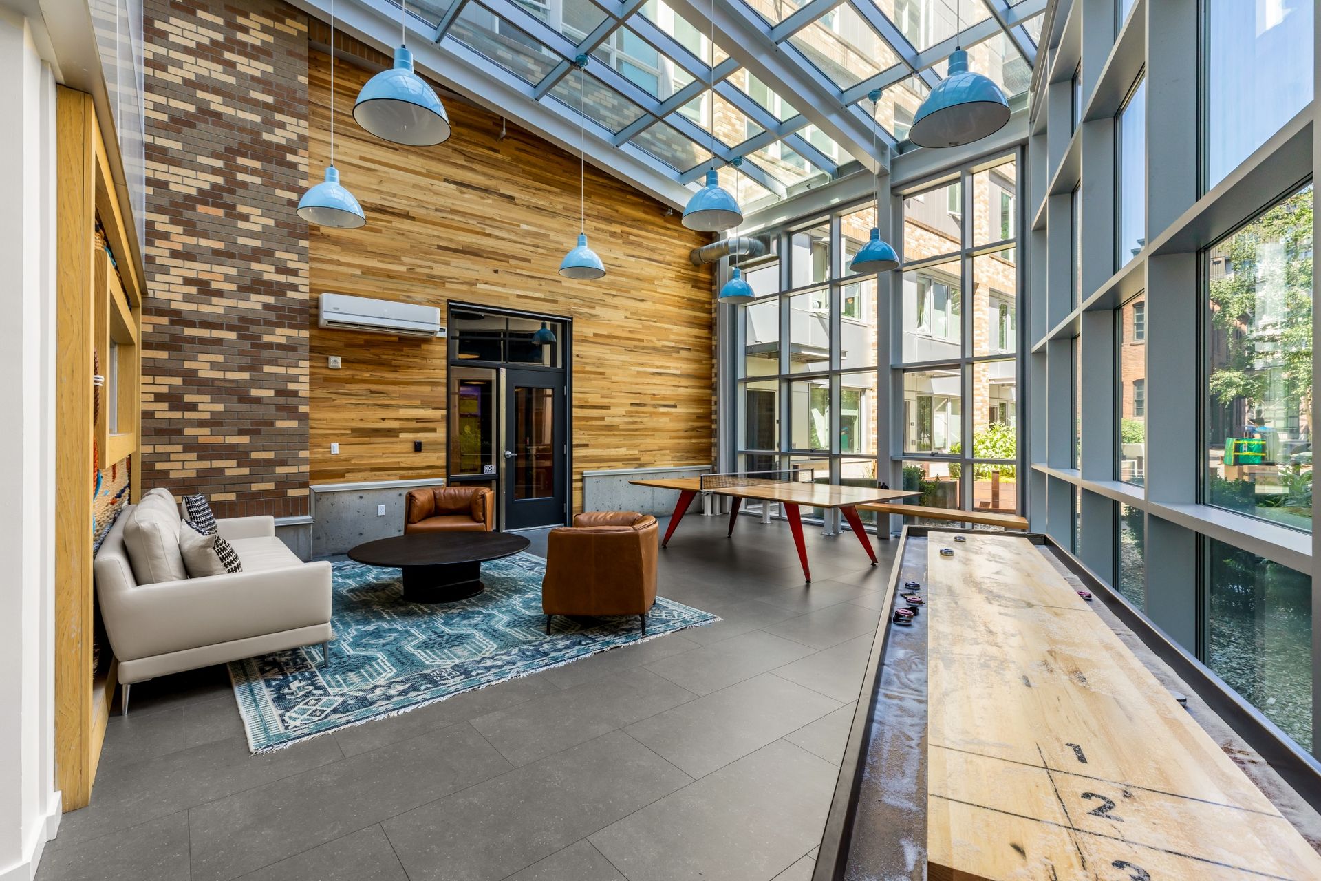 Lounge area with a glass roof, brick and wood walls, and seating. Includes a sofa, chairs, and a shuffleboard table.