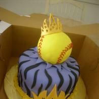 Yellow softball on purple-and-black cake with a yellow flame topper in a cardboard box