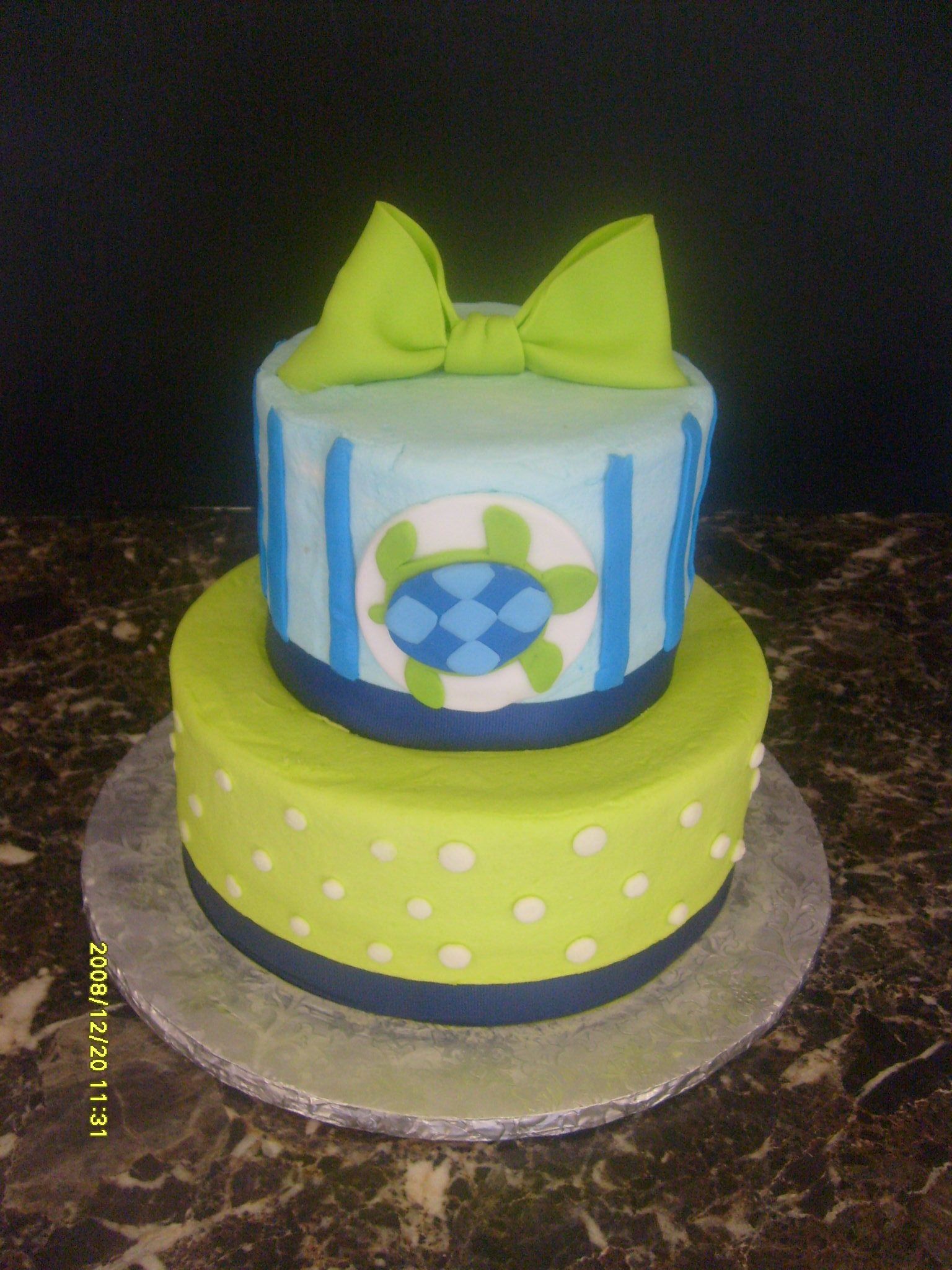 Two-tier birthday cake with lime green and blue icing and a green bow topper on a silver board