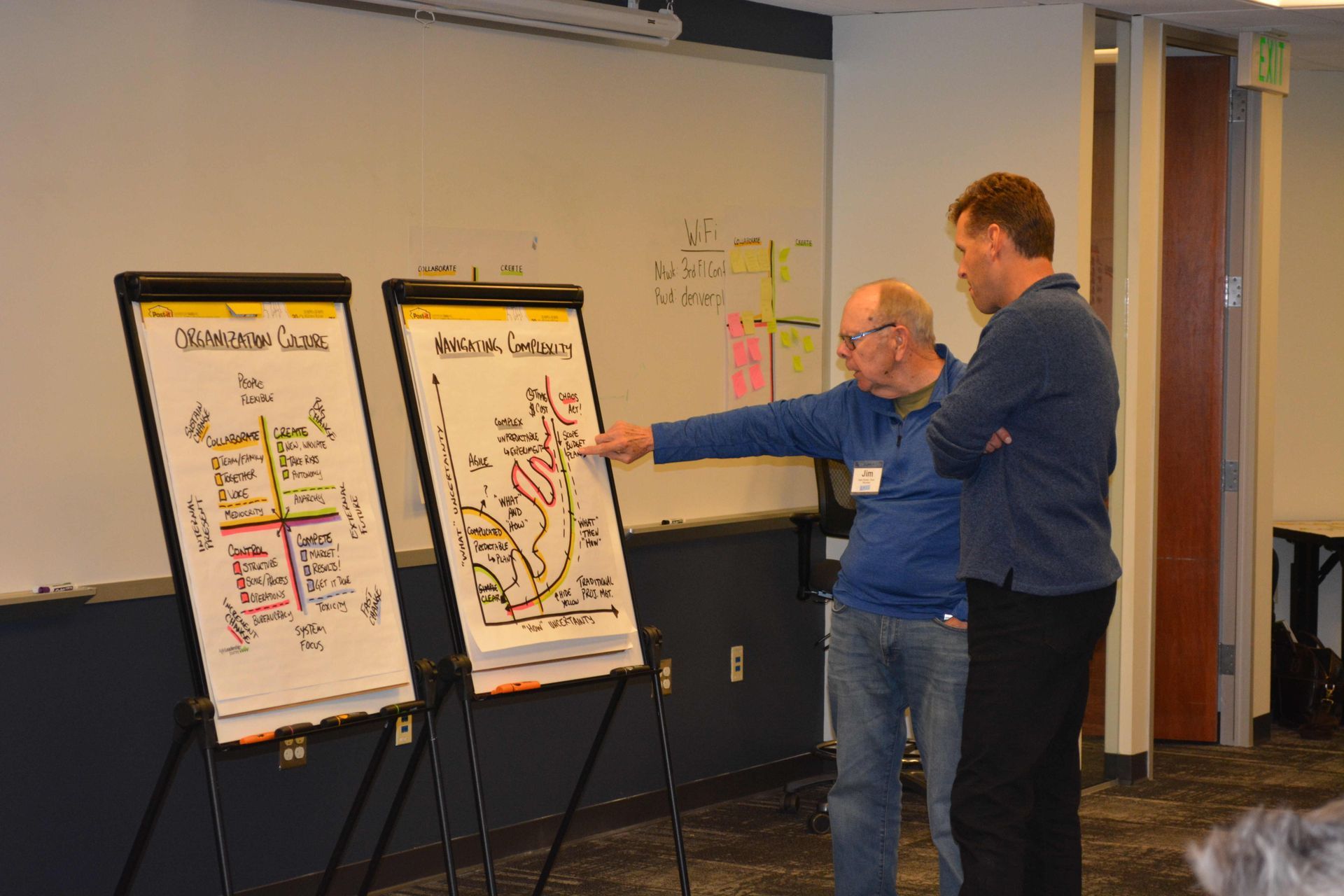 Two men are standing in front of a white board and pointing at it.