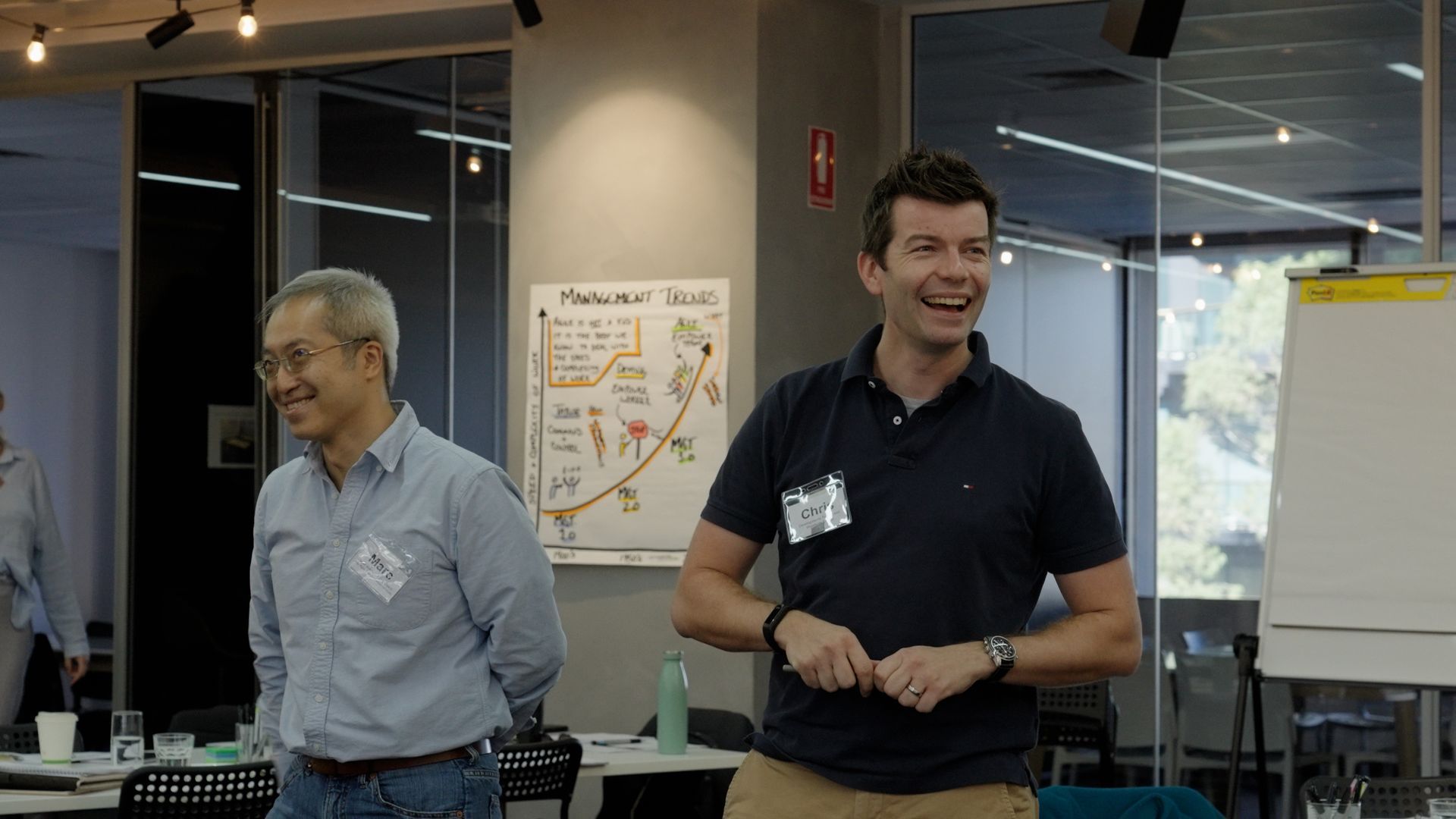 Two men are standing next to each other in a workshop room and smiling.