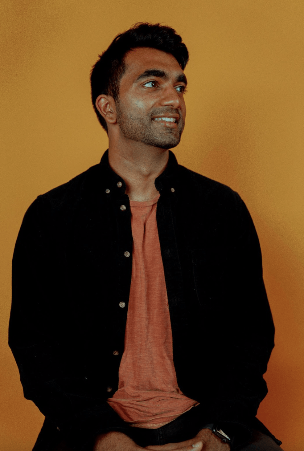 A photo of Ameet Kallaracki, a dark-skinned man. He is wearing a black button down shirt, open to reveal an orange shirt underneath. He gazing off to the right and smiling slightly.