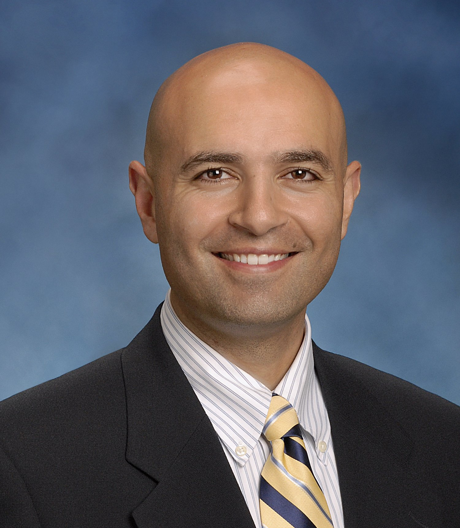 Man with shaved head smiles, wearing a suit, tie, and striped shirt against a blue backdrop.