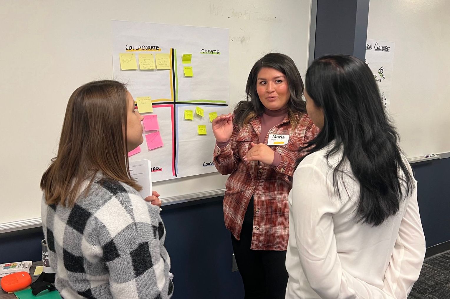 Three women are standing in front of a whiteboard talking to each other.