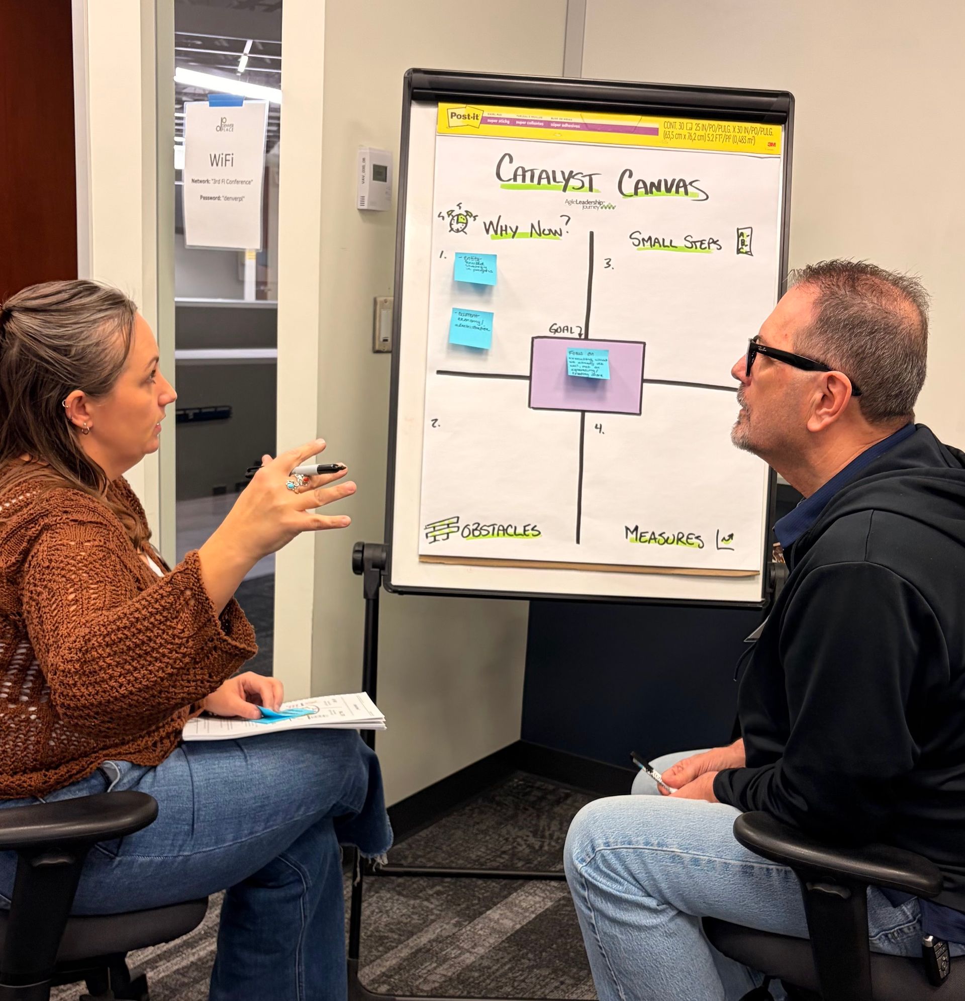 Woman pointing at a whiteboard with sticky notes, talking to a man. Office setting, focus on discussion.