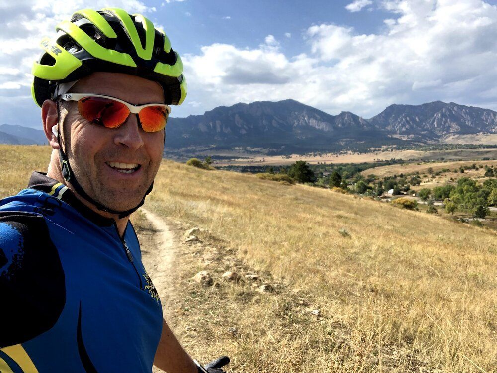 Pete taking a selfie, wearing a bike helmet and sunglasses, a mountain range is prominent in the background